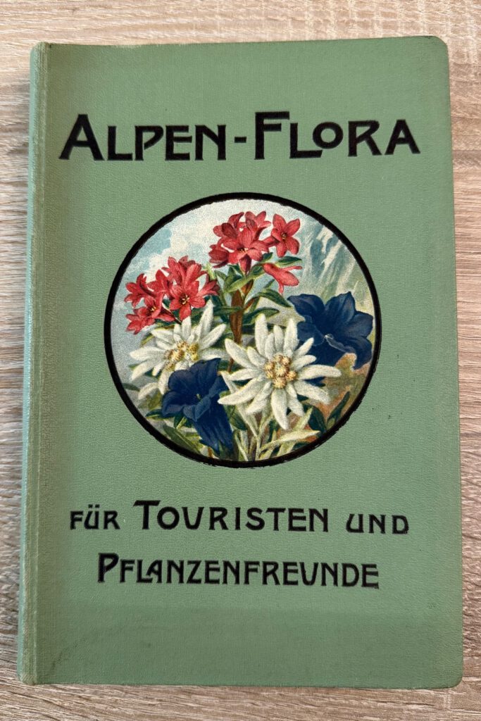 Grünes Buch mit dem Titel 'Alpen-Flora für Touristen und Pflanzenfreunde' und einem runden Bild mit roten, weißen und blauen Alpenblumen auf dem Einband.
