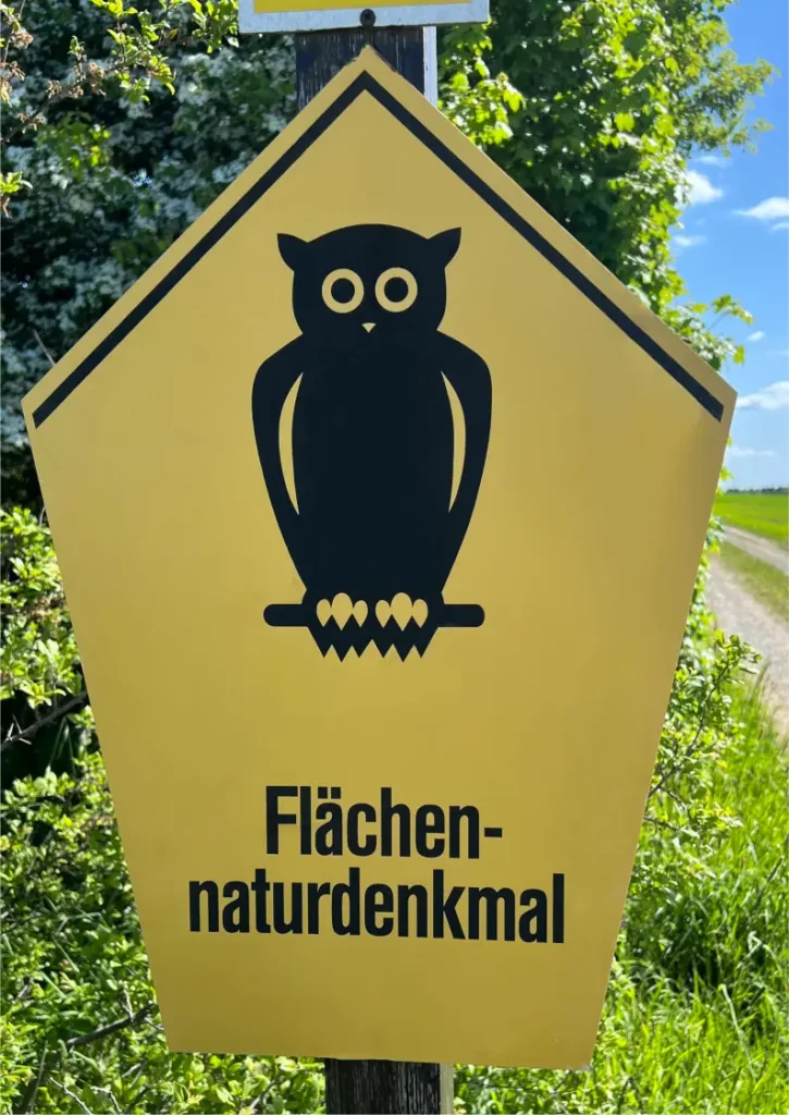 Gelbes Schild mit schwarzer Eulen-Silhouette und der Aufschrift 'Flächennaturdenkmal' vor grünem Hintergrund